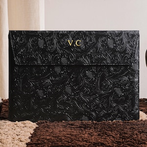 May include: Black leather laptop sleeve with embossed floral pattern and gold initials "V.C." on the flap. The sleeve is closed and sits on a textured brown and beige surface. A brown leather bag is visible in the background.