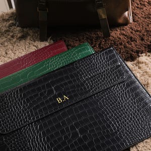 May include: Black, green, and burgundy leather laptop sleeves with a crocodile skin texture. The black sleeve has gold initials "B.A." on the front. A brown leather bag is in the background.