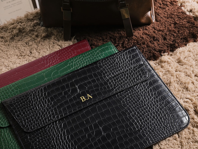 May include: Black, green, and burgundy leather laptop sleeves with a crocodile skin texture. The black sleeve has gold initials "B.A." on the front. A brown leather bag is in the background.