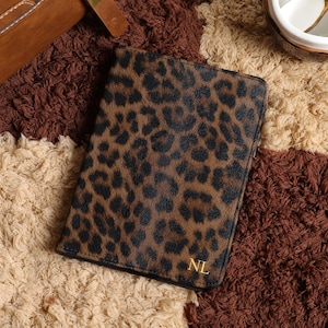 May include: A brown and black leopard print passport holder with gold initials "NL". The passport holder is on a multi-colored brown, tan, and cream textured surface. A brown leather bag is in the background.
