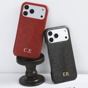 Pode incluir: Duas capas de telemóvel personalizadas. Uma é vermelha com as iniciais "C.E." e a outra é preta com as iniciais "E.R." Ambas as capas têm um padrão texturizado e um módulo de câmara com três lentes e um flash.