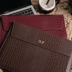 May include: Two laptop sleeves with a faux crocodile skin texture. One is a deep burgundy color, and the other is a rich brown. The brown sleeve has the gold initials "D.P." on the front. A laptop and a mug are in the background.