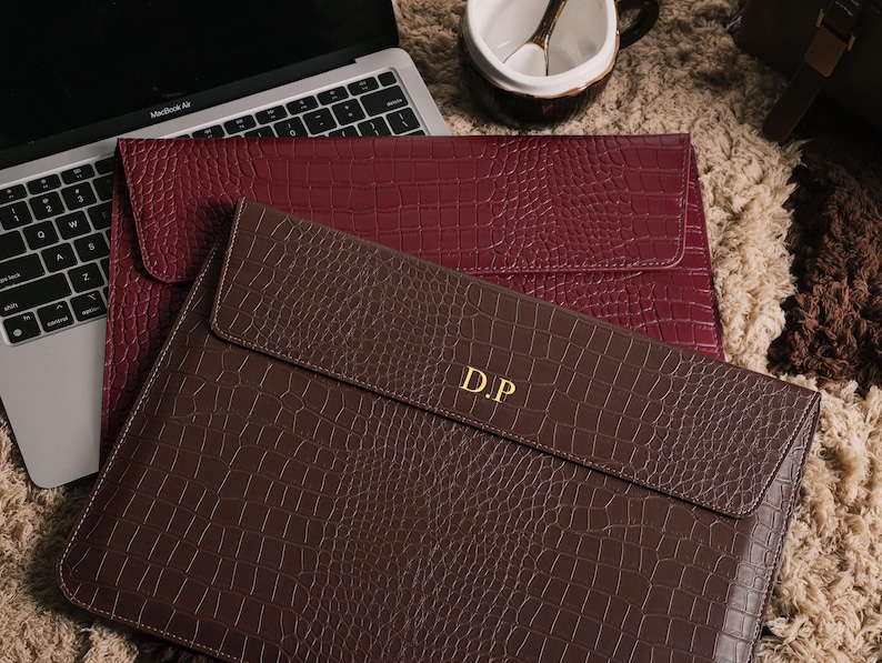 May include: Two laptop sleeves with a faux crocodile skin texture. One is a deep burgundy color, and the other is a rich brown. The brown sleeve has the gold initials "D.P." on the front. A laptop and a mug are in the background.