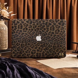 May include: A laptop skin featuring a brown and black leopard print pattern. The skin covers the exterior of a laptop, displaying a centered silver Apple logo and the gold-colored initials ES printed in the bottom right corner of the cover.