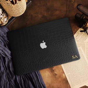 May include: A black laptop cover with a textured crocodile skin pattern, featuring a silver Apple logo in the center and the gold-colored initials SA printed in the bottom right corner. The cover is designed to fit over the exterior of a laptop.