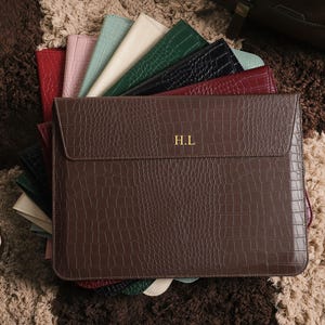 Op de afbeelding: Een bruine, imitatie-krokodil laptophoes met de initialen "H.L" in goud. Verschillende andere gekleurde hoezen, waaronder rood, roze en groen, zijn erachter uitgespreid. De items liggen op een getextureerd, veelkleurig tapijt.