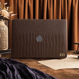 May include: A dark brown laptop skin with a textured crocodile skin pattern. The skin features a central cutout for the Apple logo and the gold-colored initials ED printed in the bottom right corner, providing a custom look for the device.