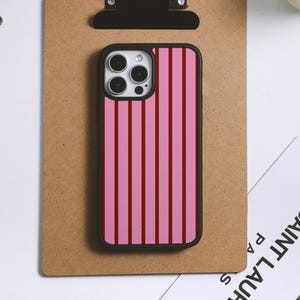 May include: A smartphone case with a pink and red striped pattern. The case has a black border and is placed on a brown clipboard. The phone case is rectangular and has a camera cutout.