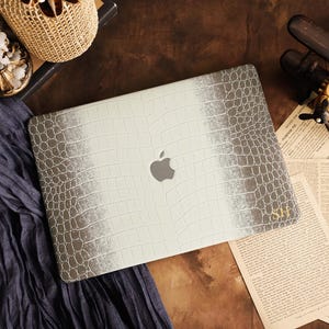Peut inclure: Un autocollant pour ordinateur portable présentant un motif de texture peau de crocodile blanc et gris avec un effet dégradé. Le centre affiche une découpe du logo Apple argenté, et le coin inférieur droit porte les initiales SH dorées.