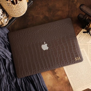 May include: A dark brown laptop cover with a textured crocodile skin pattern. The cover features a central cutout for the Apple logo and the gold-colored initials SH printed in the bottom right corner, providing a sleek and protective aesthetic.