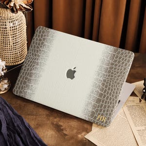 K&ouml;nnte beinhalten: Eine Laptop-H&uuml;lle mit einer wei&szlig;en und grauen Krokodilleder-Textur und einem Verlaufseffekt. In der Mitte befindet sich eine Aussparung f&uuml;r das Apple-Logo, und in der unteren rechten Ecke sind die goldenen Initialen HR aufgedruckt.