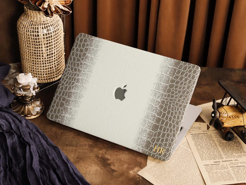 K&ouml;nnte beinhalten: Eine Laptop-H&uuml;lle mit einer wei&szlig;en und grauen Krokodilleder-Textur und einem Verlaufseffekt. In der Mitte befindet sich eine Aussparung f&uuml;r das Apple-Logo, und in der unteren rechten Ecke sind die goldenen Initialen HR aufgedruckt.