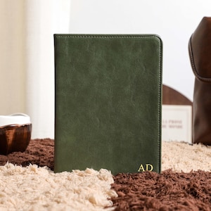 May include: A dark green leather journal with the initials "AD" in gold lettering. The journal is on a textured surface with a brown leather backpack in the background. A small ceramic bowl is also visible.