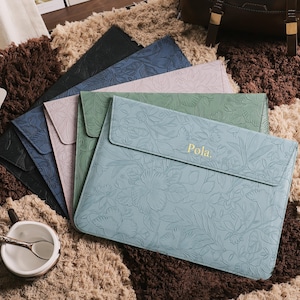 May include: A collection of laptop sleeves in various colors, including black, blue, green, and pink. Each sleeve features a floral embossed design and a magnetic flap closure. The light blue sleeve has the name "Pola" in gold lettering.