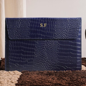 May include: A navy blue leather laptop sleeve with a crocodile skin texture. The flap closure is embossed with the gold initials "S.F." The sleeve is on a brown and beige patterned rug.