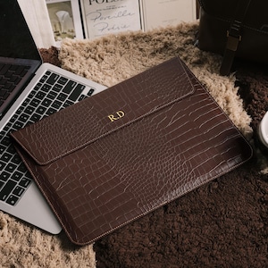 May include: A brown leather laptop sleeve with a crocodile texture and the initials "R.D" in gold. The sleeve is closed and lies on a textured surface next to a laptop. The overall theme is business and office accessories.