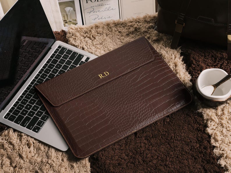 May include: A brown leather laptop sleeve with a crocodile texture and the initials "R.D" in gold. The sleeve is closed and lies on a textured surface next to a laptop. The overall theme is business and office accessories.