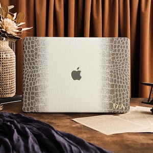 May include: A laptop skin featuring a white and grey crocodile skin texture pattern with a gradient effect. The center displays a cutout for the Apple logo, and the bottom right corner features the gold-colored initials D.A. printed on the surface.