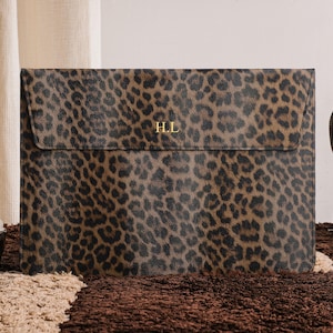 Puede incluir: Una funda para portátil con estampado de leopardo. La funda es marrón y negra con las iniciales doradas "H.L." en la parte delantera. En el fondo se ve un bolso de cuero marrón oscuro.