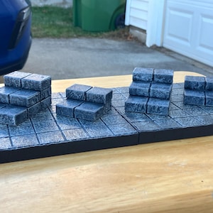 May include: A tabletop game terrain set featuring modular stone-textured tiles and stair-like structures. The tiles are gray with a grid pattern, and the stairs are made of similar material. The set is designed for tabletop gaming.