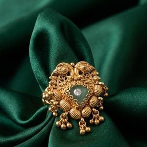 Könnte beinhalten: Ein goldfarbener Ring mit einem kunstvollen Design, das einen zentralen grünen Stein und einen kleineren, klaren Stein aufweist. Der Ring hat aufwendige Details, darunter kleine Perlenakzente und hängende Elemente. Der Ring wird auf einem dunkelgrünen Satinstoff präsentiert.