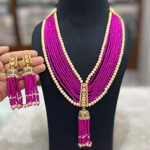 May include: A vibrant magenta beaded necklace and matching earrings set. The necklace features multiple strands of small beads, accented with white pearls. The earrings have a gold-colored top with magenta bead tassels. The jewelry is displayed on a black mannequin.