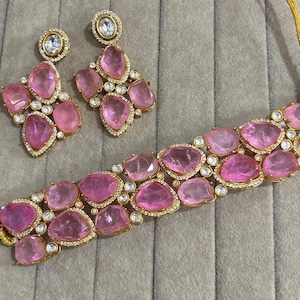Könnte beinhalten: Ein goldfarbenes Schmuckset mit einer Halskette und passenden Ohrringen. Das Set ist mit großen, rosa, unregelmäßig geformten Steinen und kleinen, klaren Akzentsteinen verziert. Die Halskette und die Ohrringe sind in einem goldfarbenen Metall gefasst.