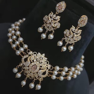 Puede incluir: Un conjunto de collar y pendientes dorados. El collar presenta tres hilos de perlas blancas y un colgante central con un dise&ntilde;o floral, cristales transparentes y una gema rosa. Los pendientes reflejan el dise&ntilde;o del colgante.