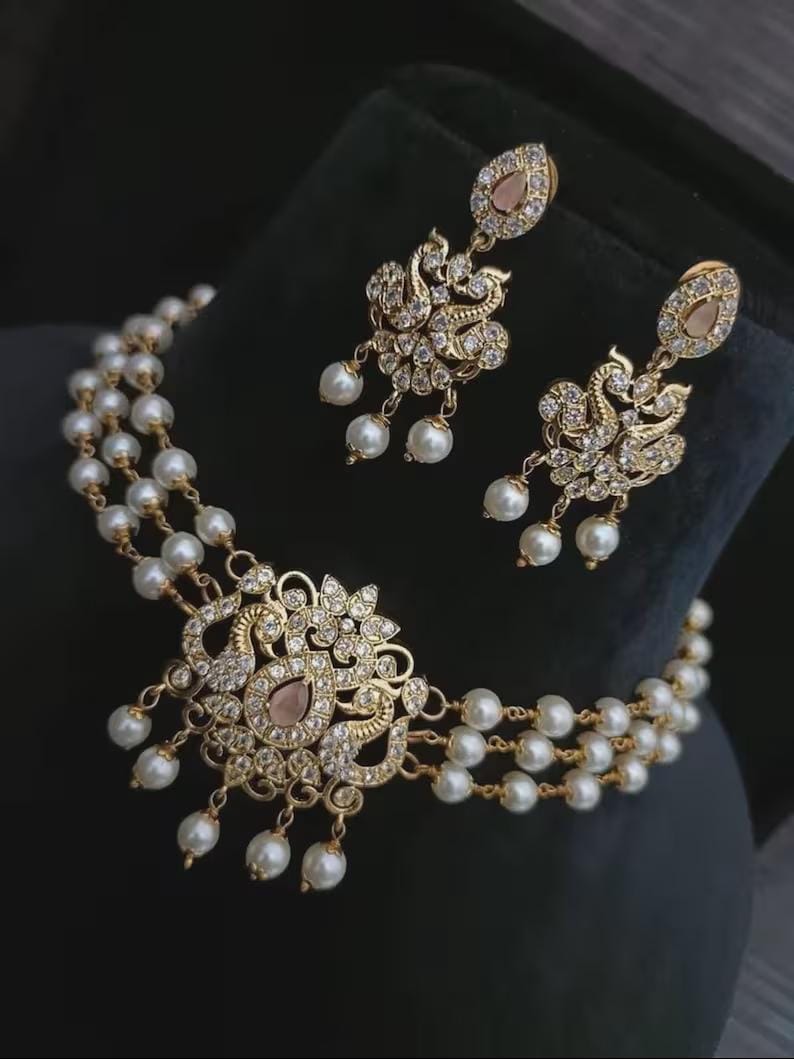 Puede incluir: Un conjunto de collar y pendientes dorados. El collar presenta tres hilos de perlas blancas y un colgante central con un dise&ntilde;o floral, cristales transparentes y una gema rosa. Los pendientes reflejan el dise&ntilde;o del colgante.
