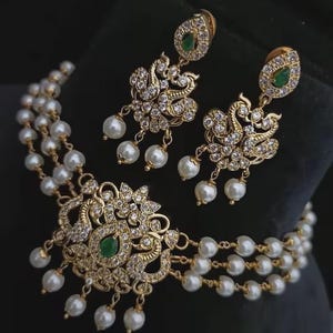 Puede incluir: Un conjunto de collar y pendientes dorados. El collar presenta un colgante central con una piedra preciosa verde y est&aacute; adornado con cristales transparentes y perlas blancas. Los pendientes a juego tienen un dise&ntilde;o similar.
