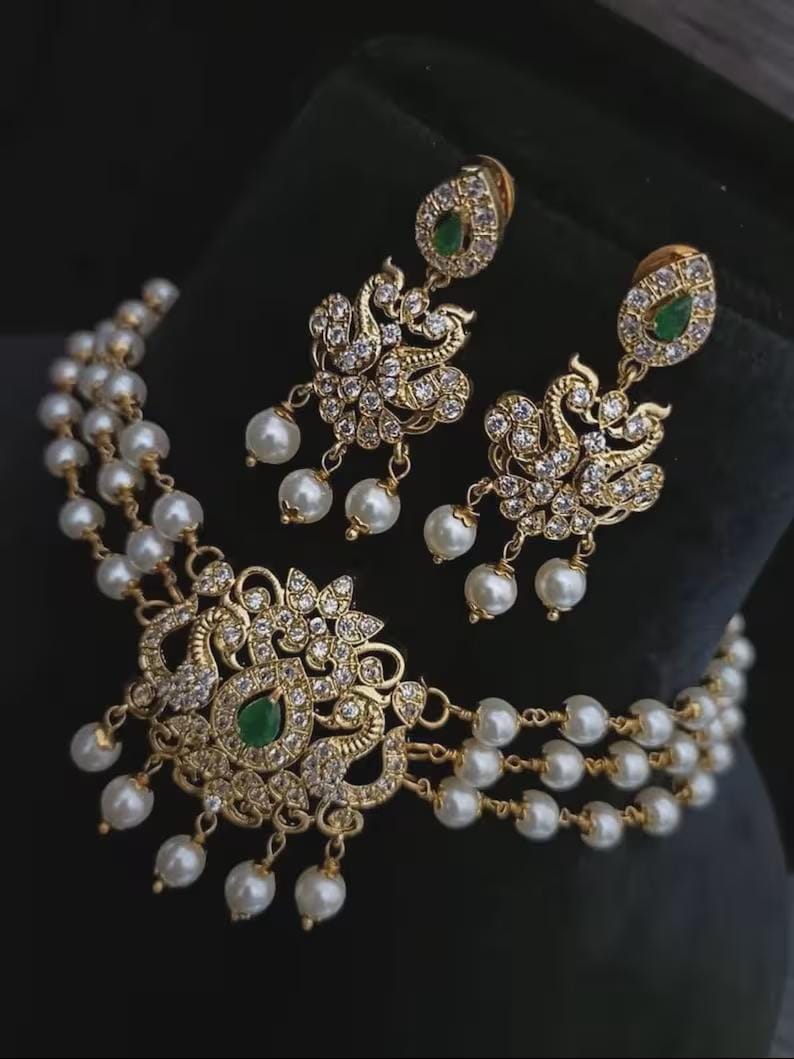 Puede incluir: Un conjunto de collar y pendientes dorados. El collar presenta un colgante central con una piedra preciosa verde y est&aacute; adornado con cristales transparentes y perlas blancas. Los pendientes a juego tienen un dise&ntilde;o similar.