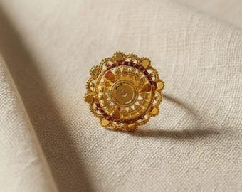 May include: A gold-colored ring with an intricate floral design. The ring features a central circular element surrounded by detailed patterns and small red accents. The ring is set against a neutral, textured background.