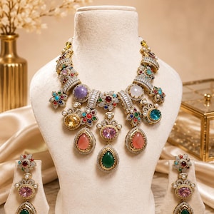 May include: A gold-toned statement necklace and matching drop earrings set featuring an array of multi-colored cabochon stones, including pink, green, blue, and yellow gems, accented with clear baguette and round crystals and small white pearls.
