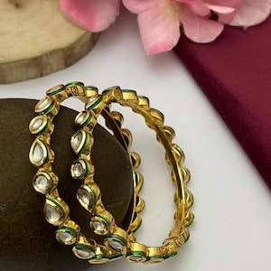 May include: Two gold bangles with teardrop-shaped clear stones and green accents. The bangles are resting on a dark brown stone, with pink flowers and a burgundy surface in the background. The bangles have a classic, elegant design.