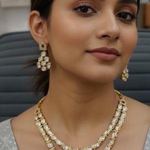 Op de afbeelding: Een goudkleurige ketting en bijpassende oorbellen set. De ketting heeft twee strengen ovale, heldere stenen gezet in goud. De oorbellen hebben een vergelijkbaar ontwerp, met meerdere heldere stenen in een gouden zetting. De persoon draagt de sieraden.