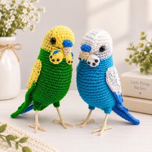 Könnte beinhalten: Zwei gehäkelte Wellensittich-Figuren. Ein Vogel hat einen gelben Kopf, einen grünen Körper und einen blauen Schwanz. Der andere hat einen weißen Kopf, einen blauen Körper und weiße Flügel. Beide haben schwarze Perlenaugen, kleine Schnäbel und helle Garnfüße.