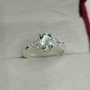 Puede incluir: Un anillo de plata con una piedra preciosa verde claro de corte ovalado en un engaste de garras, flanqueado por tres pequeñas piedras transparentes de corte marquesa a cada lado. El aro es de plata pulida y se presenta en un estuche.