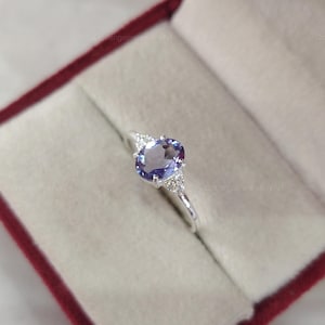 May include: A silver ring featuring an oval-cut purple gemstone as the center stone, flanked by two small clear accent stones on each side. The ring is set on a thin, polished silver band and is presented inside a red velvet jewelry box.
