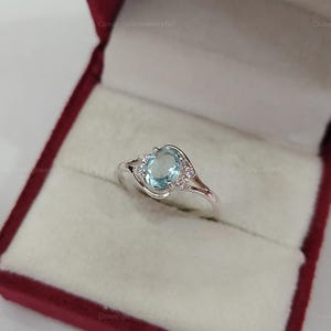 Peut inclure: Bague en argent ornée d'une pierre précieuse ovale bleu clair. La bague est sertie de petites pierres claires de chaque côté de la pierre principale. Présentée dans un écrin rouge, cette bague est élégante.