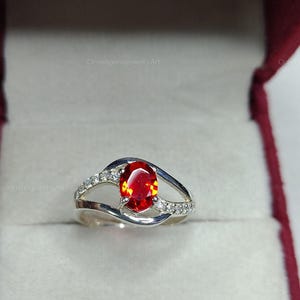 May include: A silver ring with a large, oval-cut, fiery red gemstone as the centerpiece. The band features a split design with small, clear accent stones. The ring is presented in a red jewelry box.
