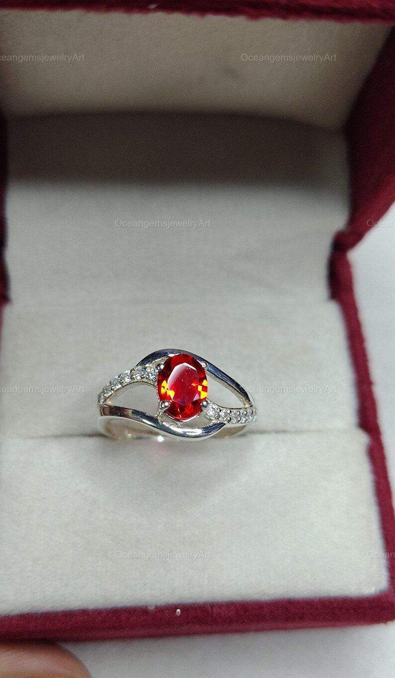 May include: A silver ring with a large, oval-cut, fiery red gemstone as the centerpiece. The band features a split design with small, clear accent stones. The ring is presented in a red jewelry box.