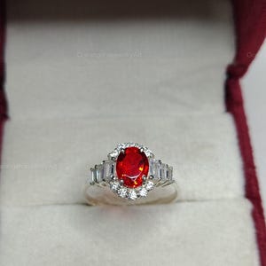 K&ouml;nnte beinhalten: Silberring mit einem gro&szlig;en, ovalen, feuerroten Edelstein in der Mitte, umgeben von kleineren wei&szlig;en Steinen und rechteckigen Akzenten. Der Ring wird in einer roten Schmuckschatulle pr&auml;sentiert.