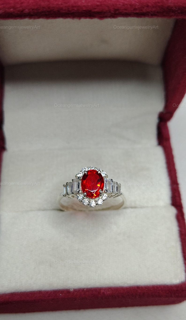 K&ouml;nnte beinhalten: Silberring mit einem gro&szlig;en, ovalen, feuerroten Edelstein in der Mitte, umgeben von kleineren wei&szlig;en Steinen und rechteckigen Akzenten. Der Ring wird in einer roten Schmuckschatulle pr&auml;sentiert.