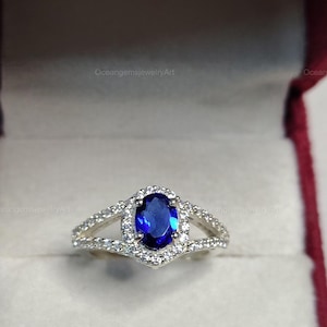 Peut inclure: Bague en argent ornée d'une grande pierre précieuse ovale bleu foncé, entourée de petites pierres claires. La bague présente un design à tige fendue et est présentée dans un écrin rouge.
