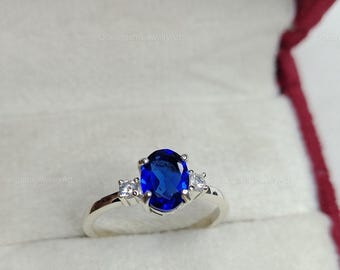 May include: A silver ring with a large, oval-cut, deep blue gemstone as the centerpiece. Two smaller, square-cut, clear gemstones flank the main stone. The ring is set against a white background inside a red jewelry box.
