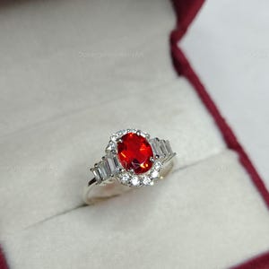K&ouml;nnte beinhalten: Ein silberner Ring mit einem gro&szlig;en, ovalen, feuerroten Edelstein, umgeben von einem Kranz aus kleinen wei&szlig;en Steinen. Der Ring hat au&szlig;erdem rechteckige wei&szlig;e Steine auf jeder Seite. Pr&auml;sentiert in einer roten Schmuckschatulle.