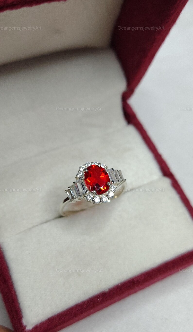 K&ouml;nnte beinhalten: Ein silberner Ring mit einem gro&szlig;en, ovalen, feuerroten Edelstein, umgeben von einem Kranz aus kleinen wei&szlig;en Steinen. Der Ring hat au&szlig;erdem rechteckige wei&szlig;e Steine auf jeder Seite. Pr&auml;sentiert in einer roten Schmuckschatulle.