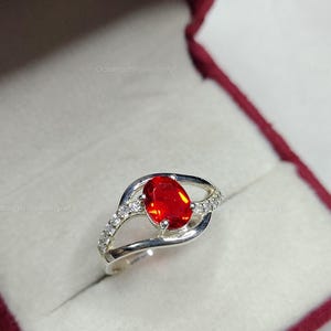 May include: A silver ring with a large, oval, bright red gemstone as the focal point. The band features a split design with small, clear accent stones. The ring is presented in a red jewelry box.