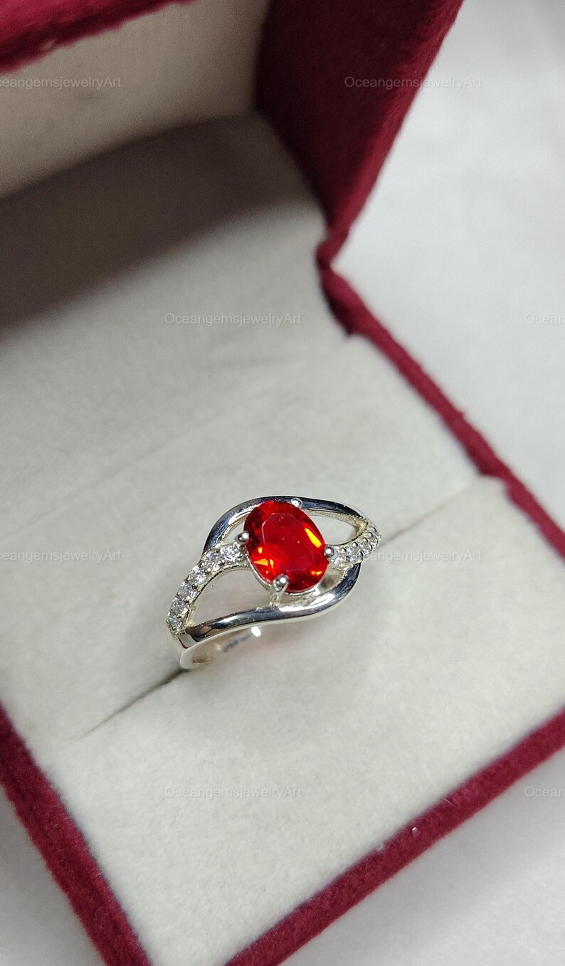 May include: A silver ring with a large, oval, bright red gemstone as the focal point. The band features a split design with small, clear accent stones. The ring is presented in a red jewelry box.