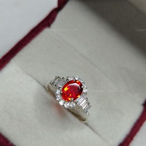 K&ouml;nnte beinhalten: Silberring mit einem gro&szlig;en, oval geschliffenen, feuerroten Edelstein als Mittelpunkt. Der Edelstein ist von einem Kranz aus kleinen, klaren Steinen umgeben. Der Ring hat auch rechteckige, klare Steine auf jeder Seite.